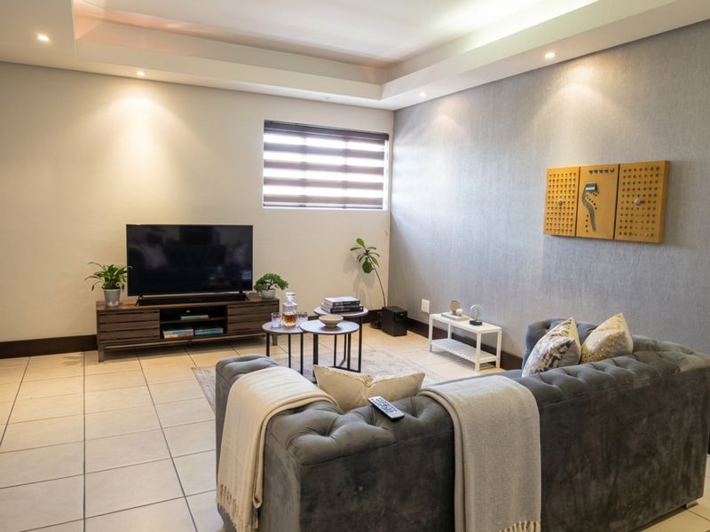 A well-lit living room featuring tiled flooring, a modern drop ceiling with recessed lighting, and neutral wall finishes with a textured accent wall.
