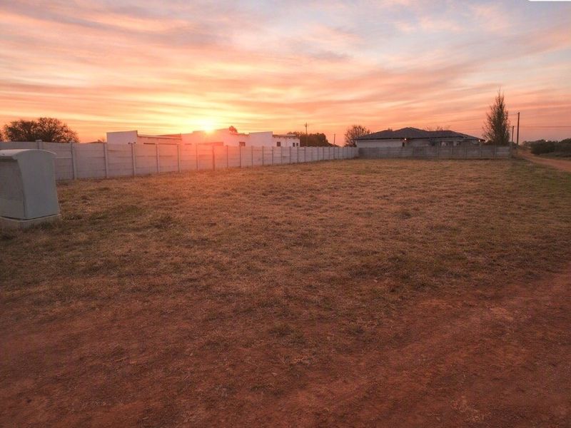 A vacant residential plot featuring a precast concrete boundary wall, an electrical utility box, and access via a dirt road at sunset.