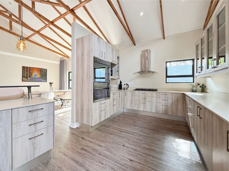 Modern open-plan kitchen featuring light wood-grain cabinetry, stone countertops, integrated appliances, and striking exposed timber roof trusses in a bright, airy space.