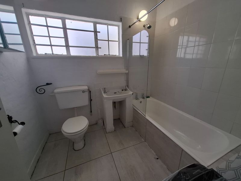 A dated bathroom featuring a pedestal basin, toilet, and a bathtub with a glass shower screen, finished with white wall tiles and tiled flooring.