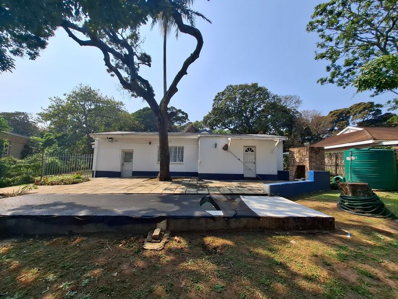 Rear exterior view featuring a paved patio, built-in braai, and a swimming pool with a damaged cover, set within a mature garden.