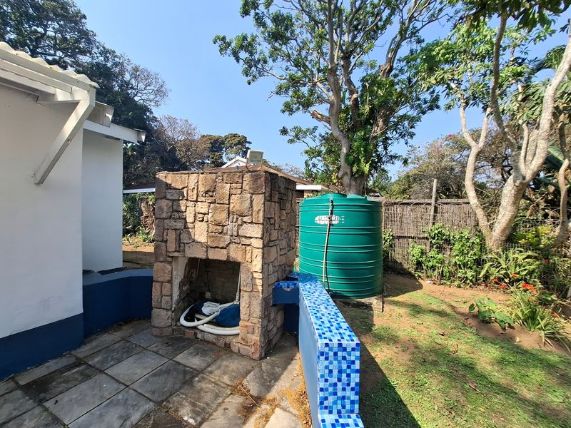 Rear garden area featuring a stone-clad outdoor fireplace, tiled patio space, and a large water storage tank set against a landscaped backdrop.