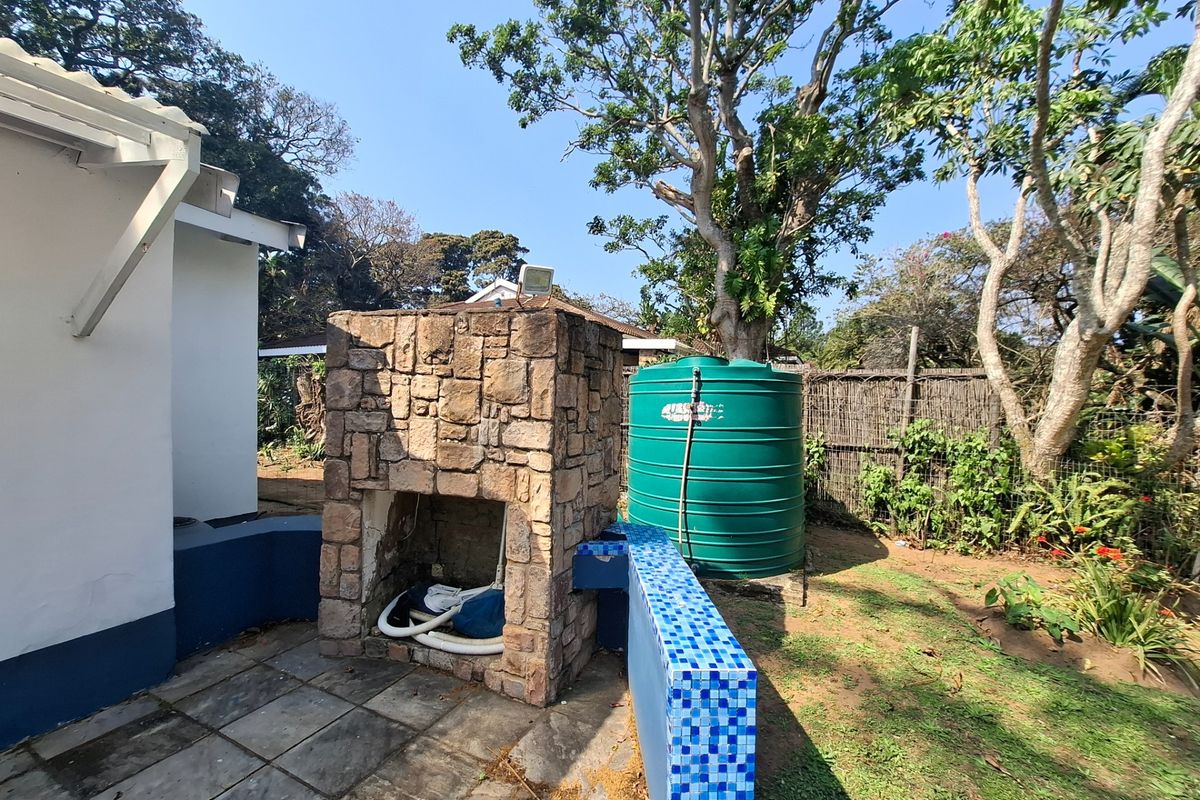 Rear garden area featuring a stone-clad outdoor fireplace, tiled patio space, and a large water storage tank set against a landscaped backdrop.