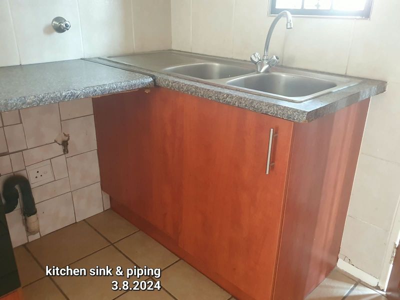 A dated kitchen area featuring a double bowl stainless steel sink, laminate countertop, and base cabinetry with visible wall tile damage and exposed plumbing.