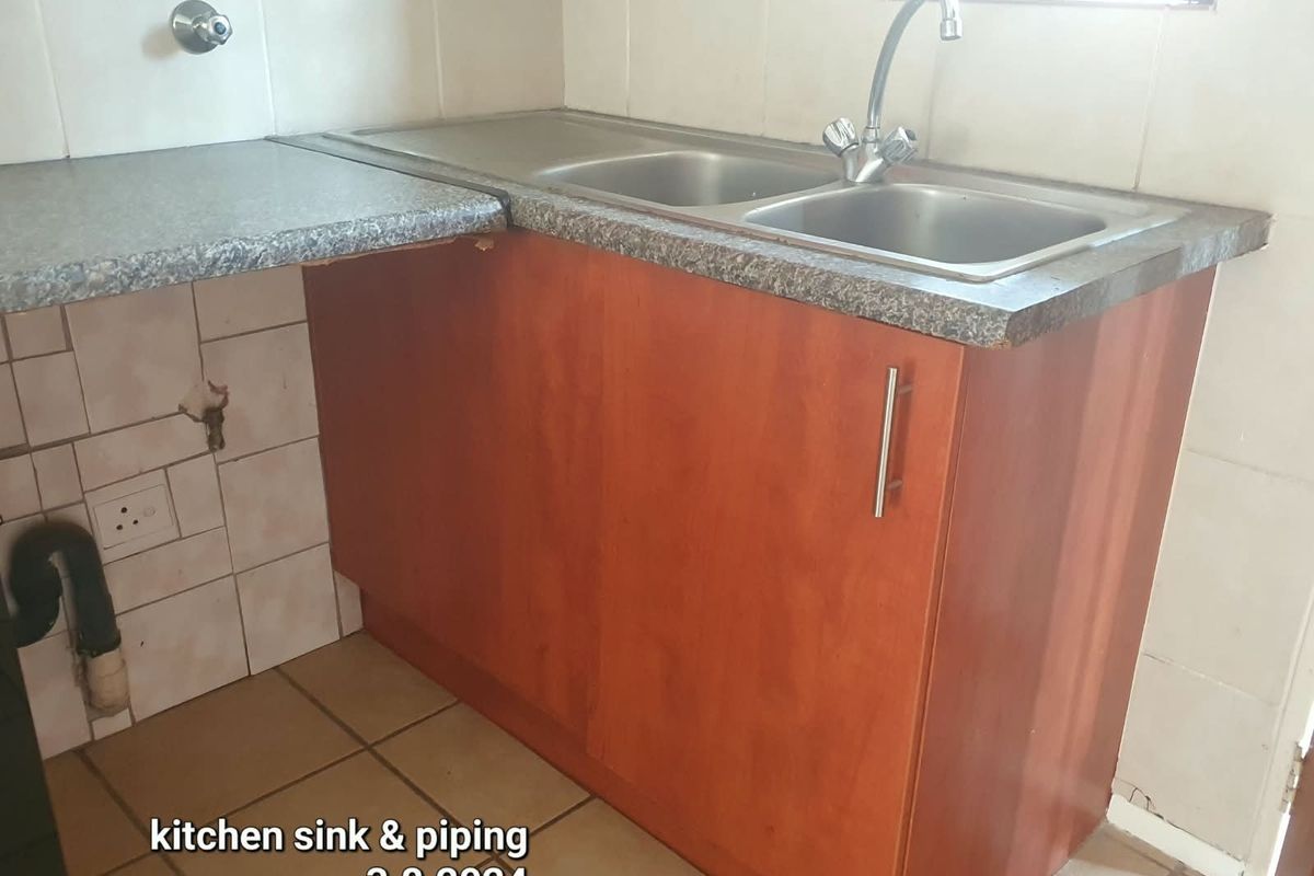 A dated kitchen area featuring a double bowl stainless steel sink, laminate countertop, and base cabinetry with visible wall tile damage and exposed plumbing.