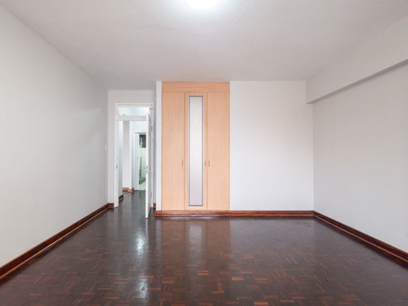 A spacious, empty bedroom featuring polished parquet flooring and a built-in wooden wardrobe with a central frosted glass panel.