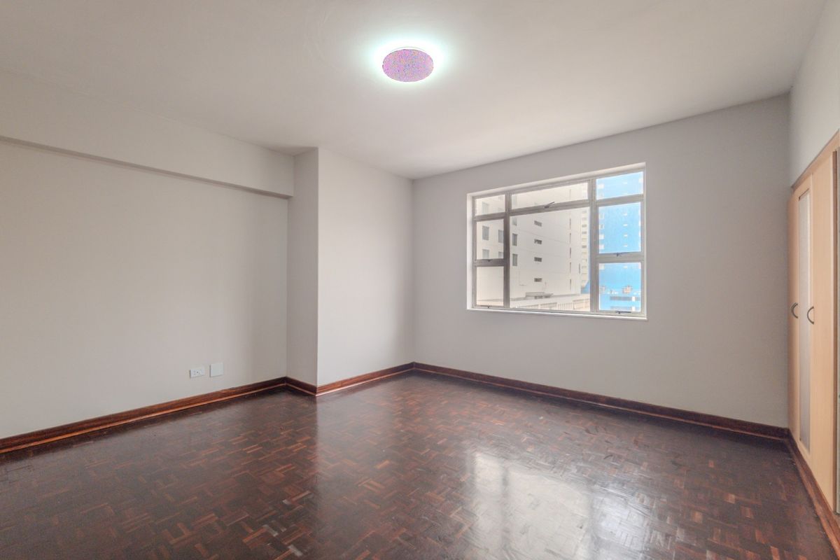 A bright, empty bedroom featuring polished parquet flooring, a built-in wardrobe, and large windows providing natural light.