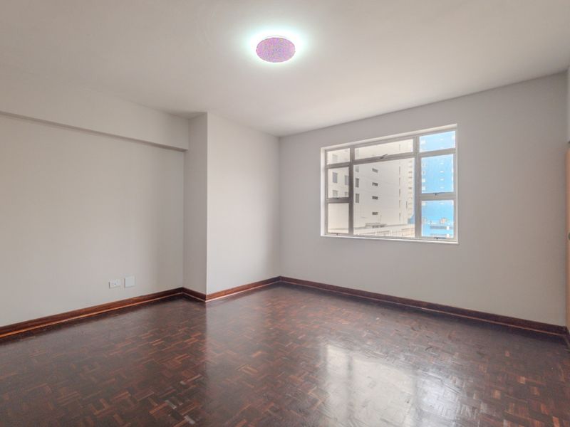 A bright, empty bedroom featuring polished parquet flooring, a built-in wardrobe, and large windows providing natural light.