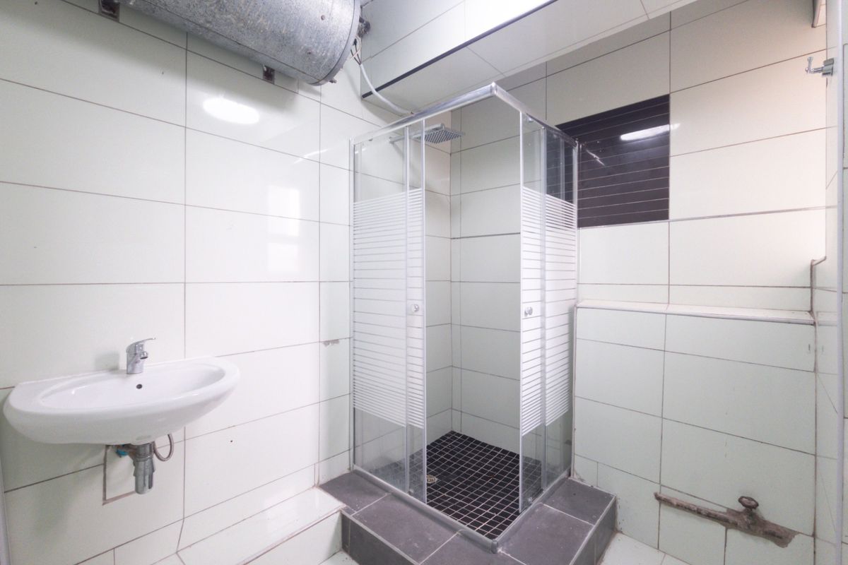 A functional bathroom featuring a shower enclosure, wall-mounted basin, and tiled finishes, currently requiring cosmetic updates and improved plumbing concealment.