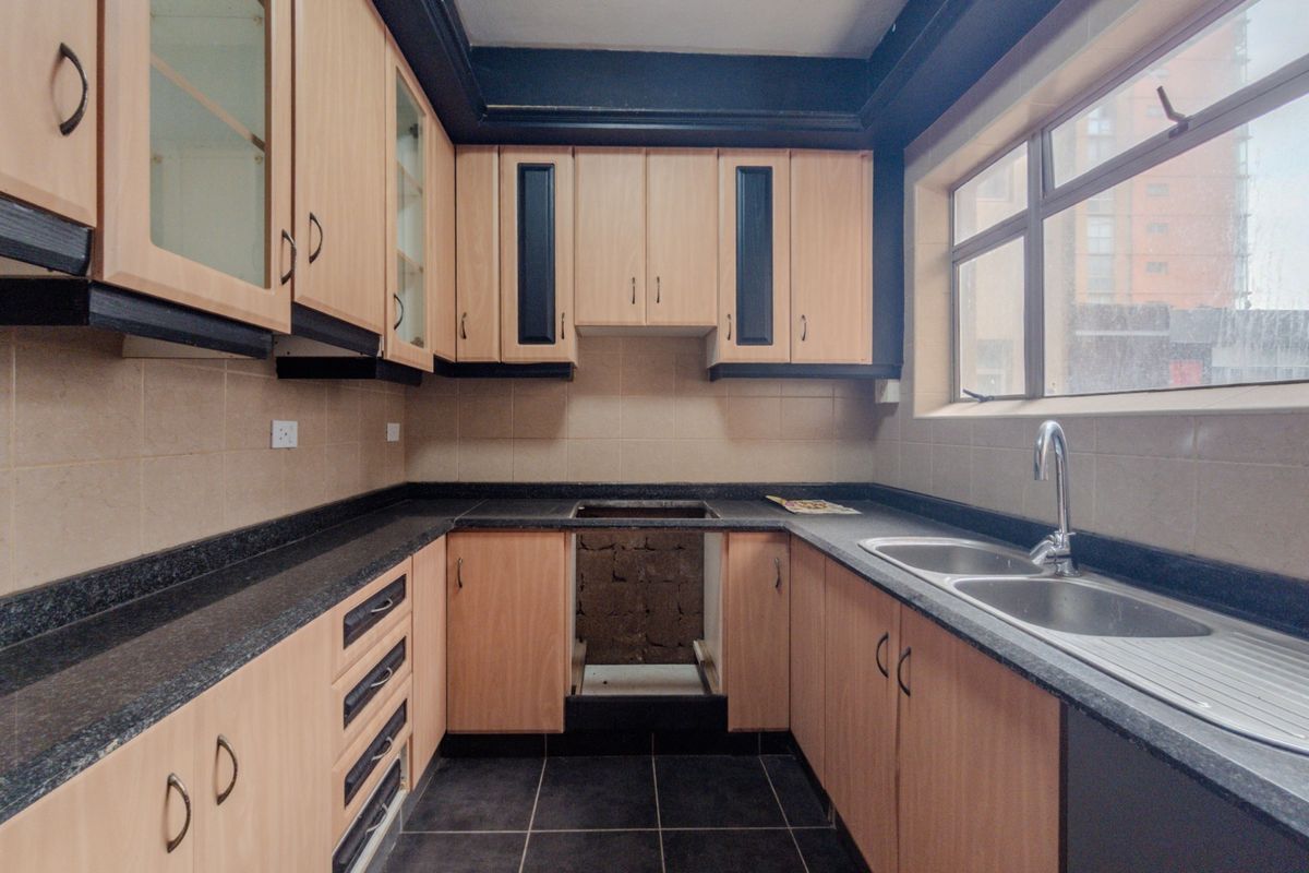A functional kitchen featuring light wood-finish cabinetry, dark granite countertops, a double stainless steel sink, and tiled finishes, awaiting a new stove installation.