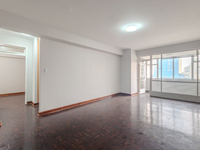 Spacious, empty living area featuring dark parquet flooring, neutral walls, and direct access to an enclosed balcony, offering a blank canvas for interior design.