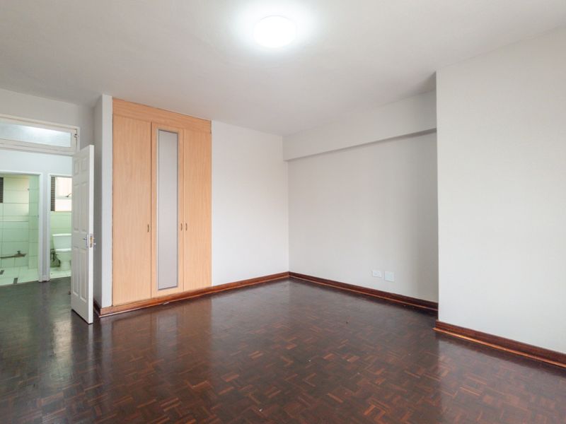 A bright, empty bedroom featuring polished parquet flooring, a built-in wardrobe with a central mirror panel, and direct access to an en-suite bathroom.