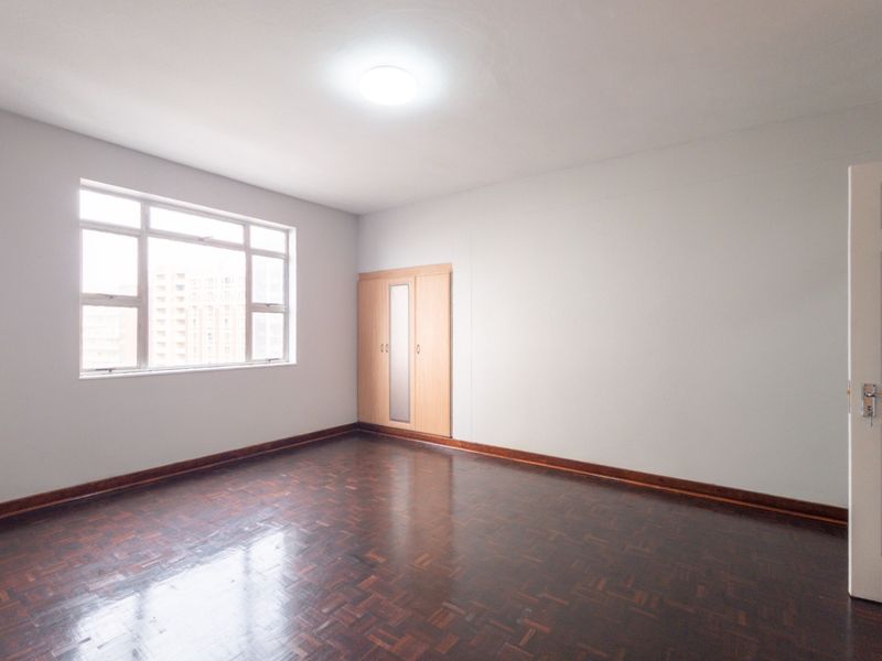 A spacious, empty bedroom featuring polished parquet flooring, a built-in wardrobe, and neutral walls, offering a clean canvas for personalization.