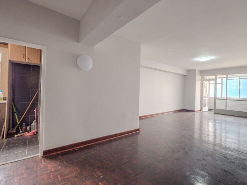 An open-plan living area featuring parquet flooring, leading to an enclosed balcony, with an adjacent kitchen area visible through the doorway.