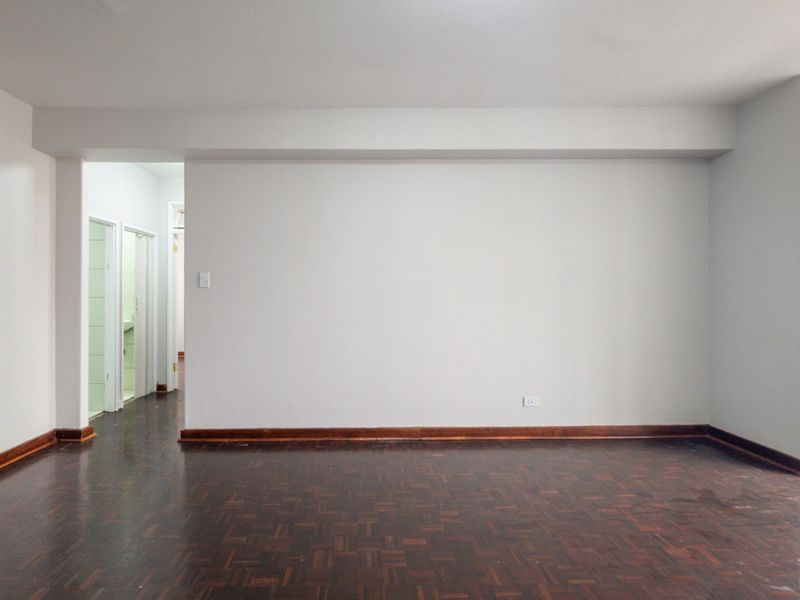 An empty living area featuring dark parquet flooring and timber skirting boards, with a clear view into the adjacent hallway.