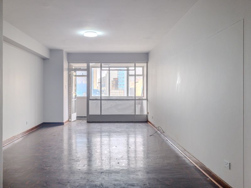 A spacious, empty living area featuring dark parquet flooring, neutral walls, and large windows providing natural light and access to an adjacent balcony.