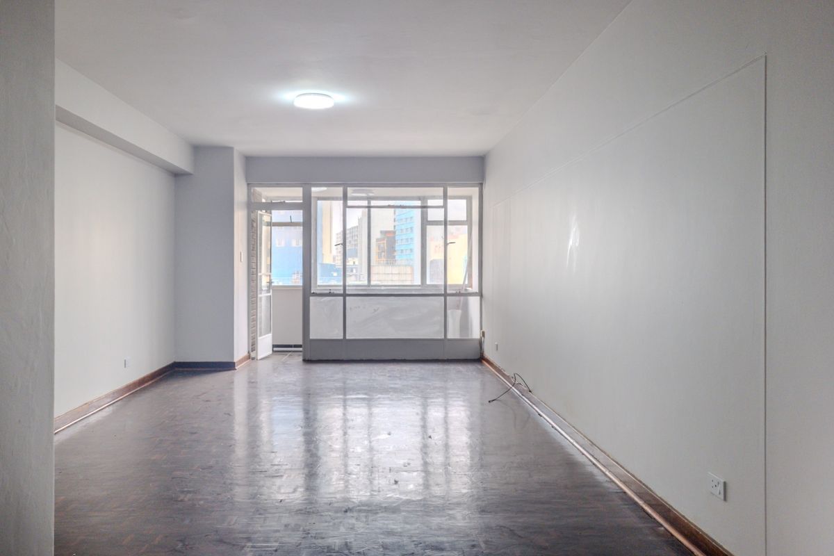 A spacious, empty living area featuring dark parquet flooring, neutral walls, and large windows providing natural light and access to an adjacent balcony.