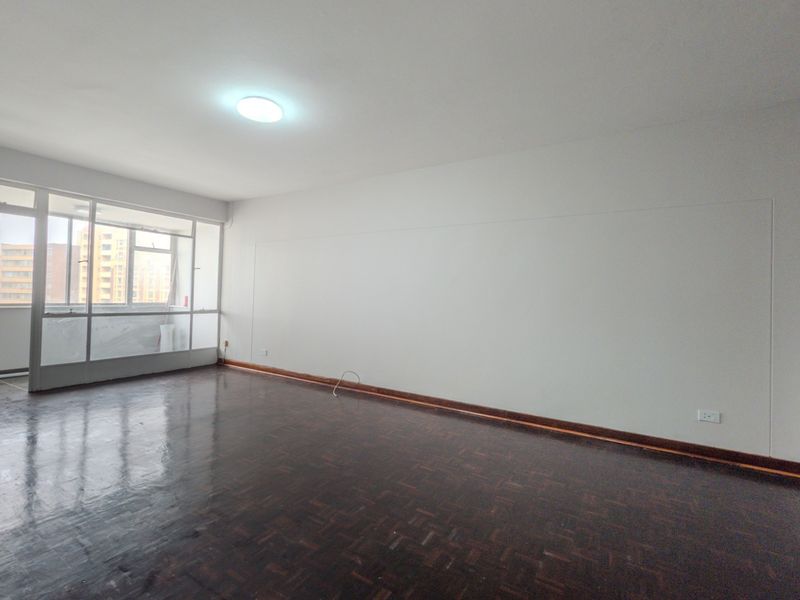 A spacious, empty living room featuring dark parquet flooring, white walls, and a large glass partition providing natural light.
