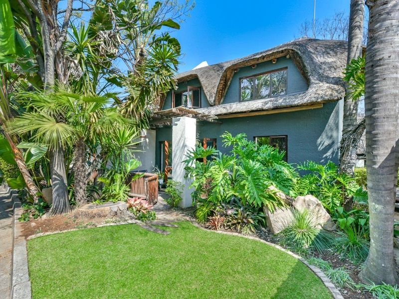 A charming townhouse exterior featuring a traditional thatched roof, a timber pergola, and a lush, well-maintained garden with a paved walkway.