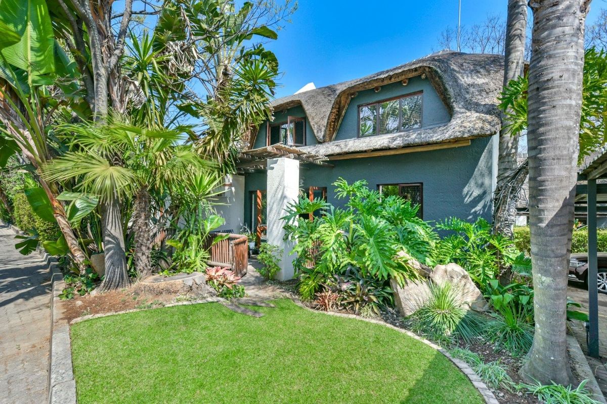 A charming townhouse exterior featuring a traditional thatched roof, a timber pergola, and a lush, well-maintained garden with a paved walkway.