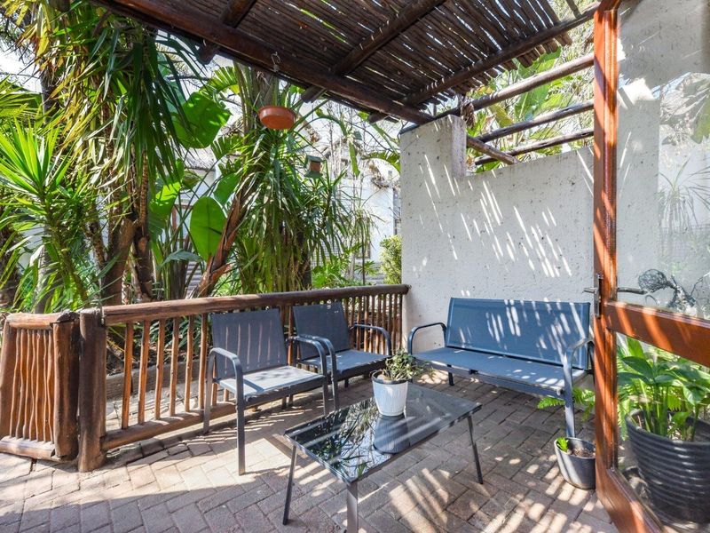 A covered outdoor patio area featuring brick paving, a timber pergola structure, and a timber railing, providing a shaded space overlooking the garden.