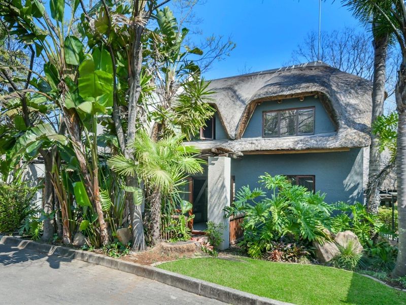 Front elevation of a two-story townhouse featuring a traditional thatched roof, mature garden landscaping, and a paved driveway entrance.