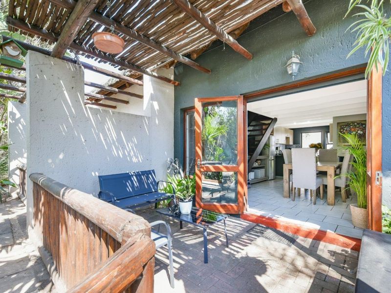 A covered patio area featuring a rustic thatched pergola, timber railing, and paved flooring, providing seamless indoor-outdoor flow through double doors into the living space.