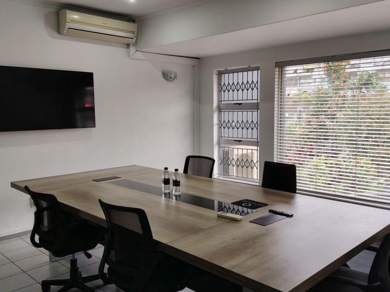 A functional office meeting room featuring tiled flooring, a wall-mounted air conditioning unit, and large windows with security gates and venetian blinds.
