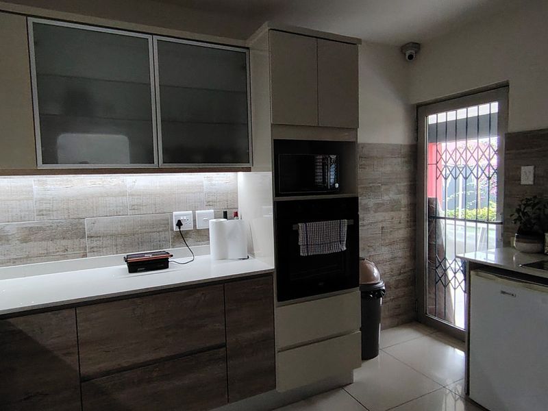 Modern kitchen featuring wood-grain cabinetry, built-in oven and microwave, stone-look tiled backsplash, and direct access to the exterior via a secure glass door.