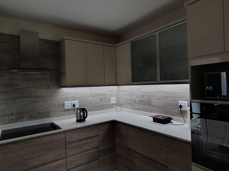 Modern kitchen featuring wood-grain cabinetry, stone countertops, integrated appliances, and stylish under-cabinet lighting with a contemporary tiled backsplash.