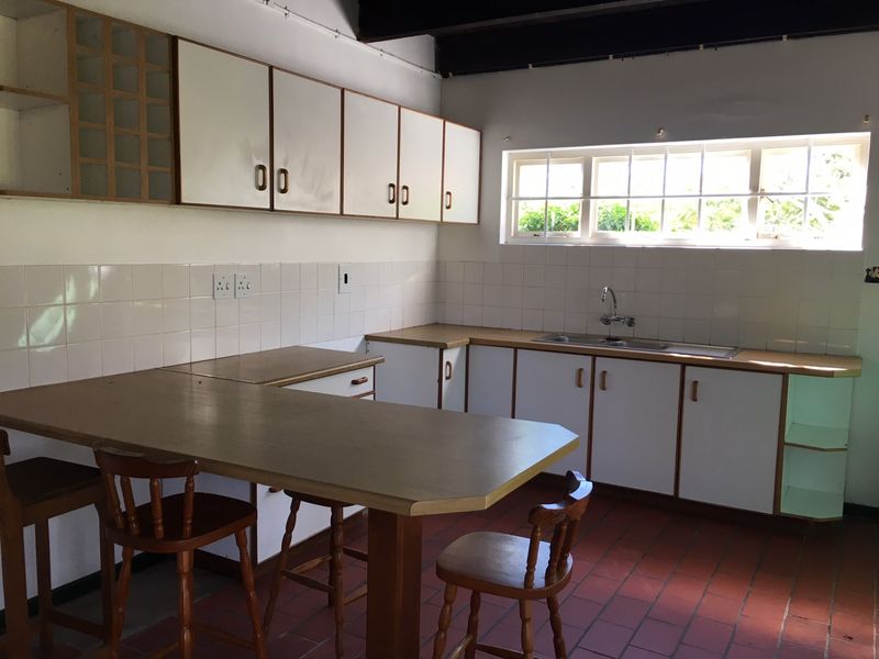A functional kitchen featuring white cabinetry, laminate countertops, a double bowl sink, and traditional terracotta floor tiles with exposed timber ceiling beams.