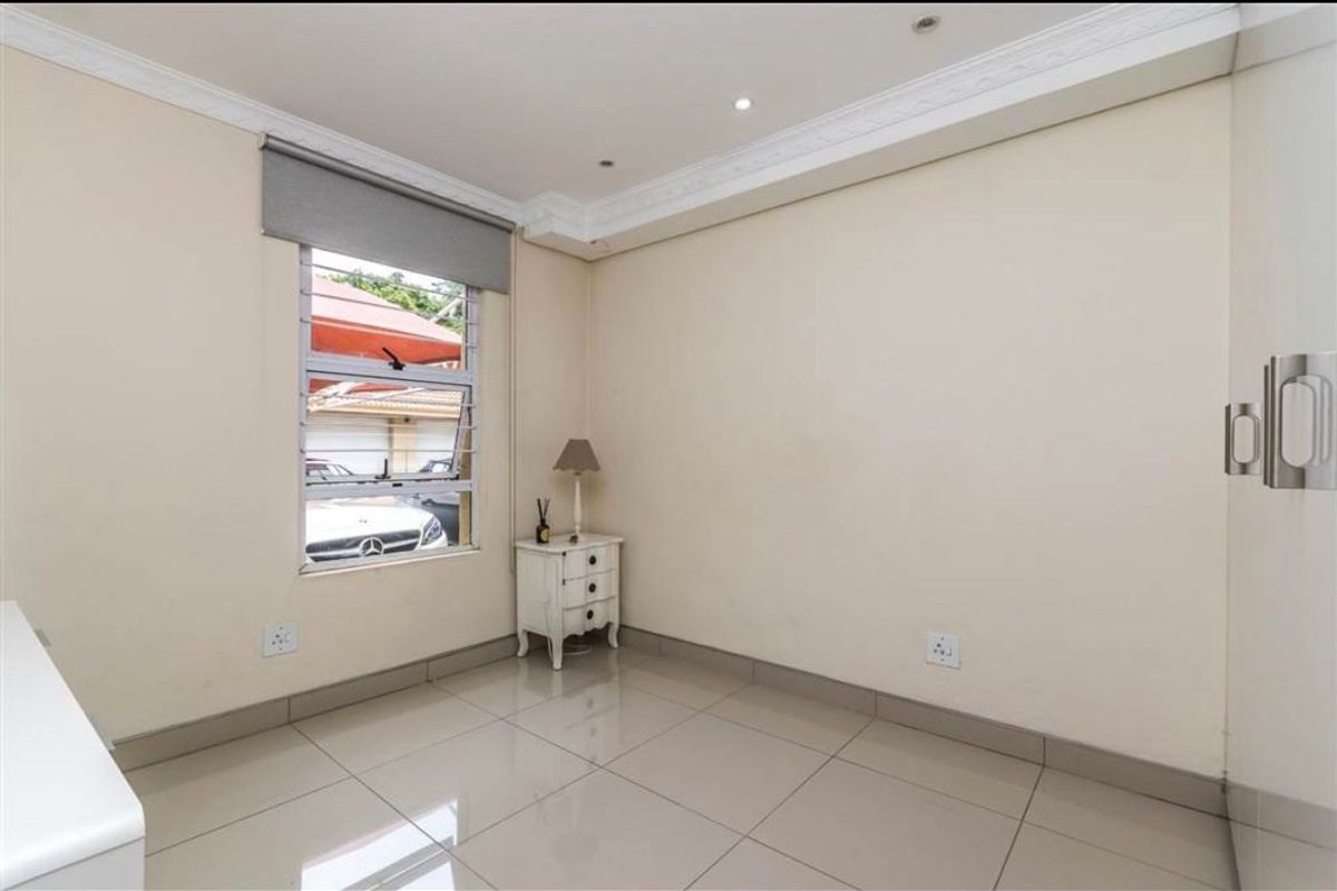 A bright, neutral bedroom featuring large floor tiles, built-in storage, and a louvered window with a roller blind.