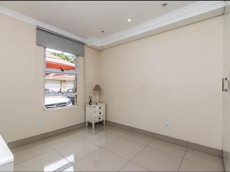 A bright, neutral bedroom featuring large floor tiles, built-in storage, and a louvered window with a roller blind.