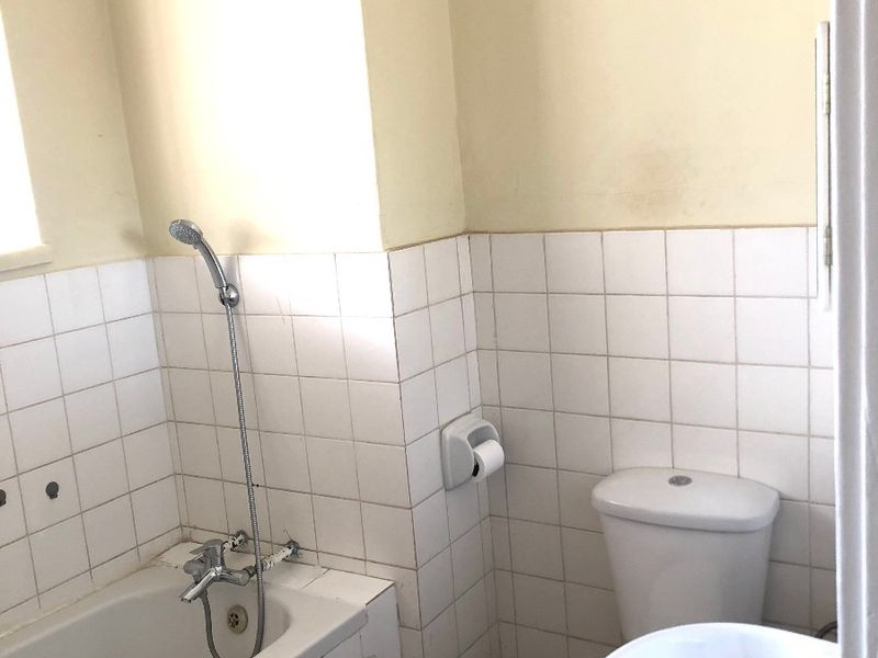 A compact bathroom featuring a bathtub with handheld shower, pedestal sink, and toilet, finished with white wall tiles and requiring cosmetic updates.