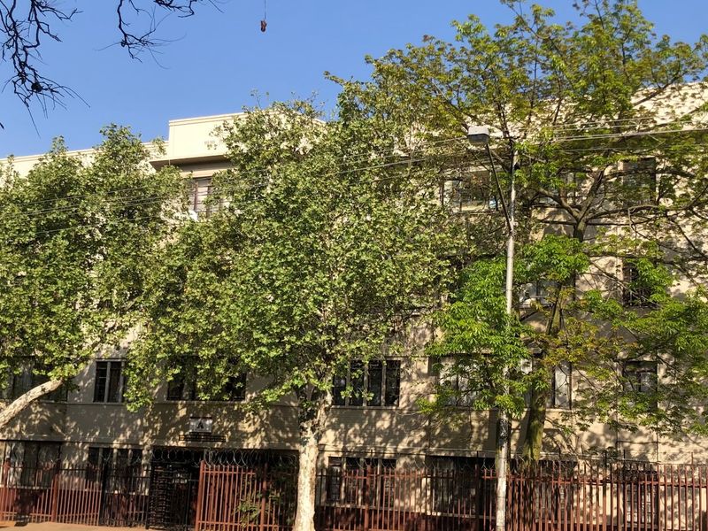 Front elevation of a multi-story apartment building featuring security fencing and mature trees along the street frontage.