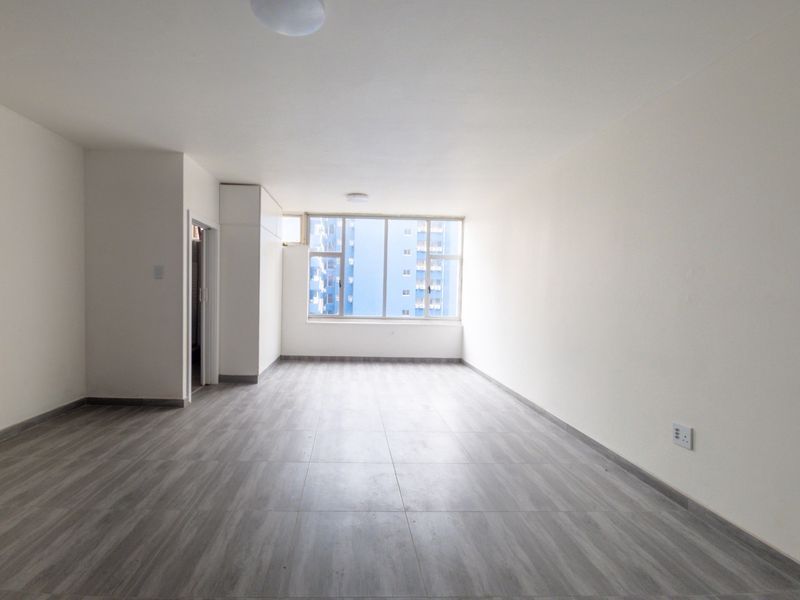 A bright, spacious, and empty living area featuring modern grey tiled flooring, neutral walls, and a large window providing natural light.