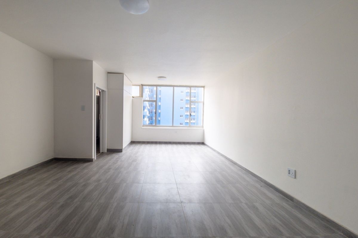 A bright, spacious, and empty living area featuring modern grey tiled flooring, neutral walls, and a large window providing natural light.