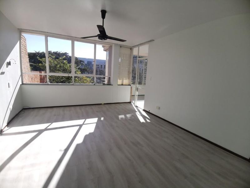 A bright, empty living room featuring modern laminate flooring, a ceiling fan, and large windows providing ample natural light.