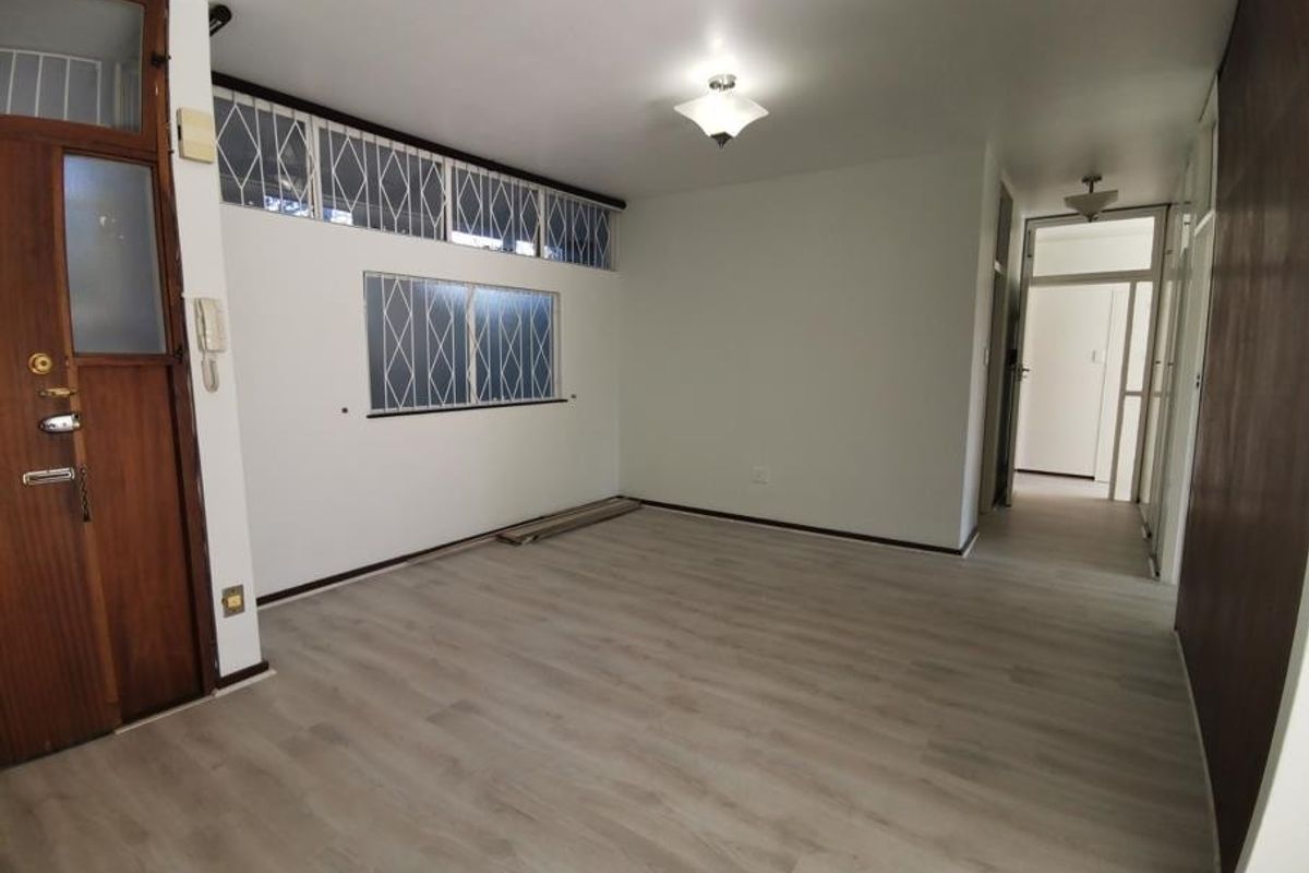 An empty living room featuring light-toned laminate flooring, security gates on windows, and an intercom system, offering a clean canvas for personalization.