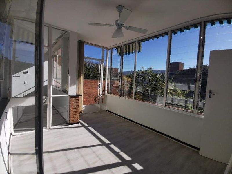 A bright, enclosed balcony or sunroom featuring laminate flooring, a ceiling fan, and expansive louver windows with views of the surrounding area.