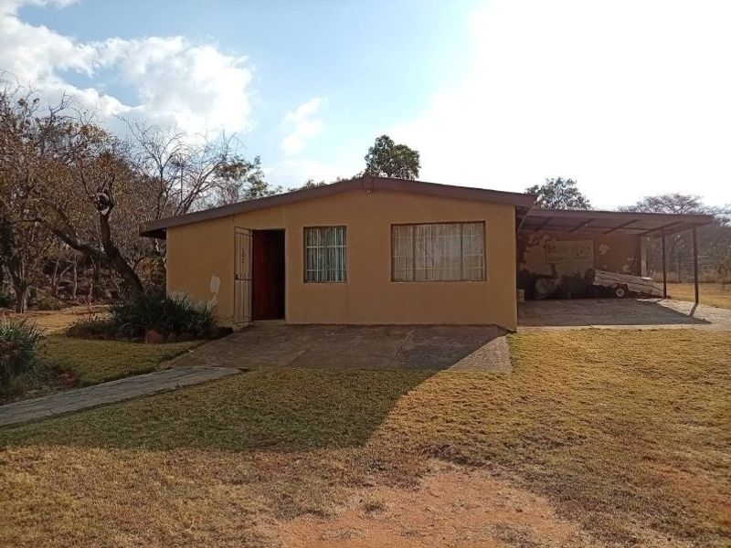 Front view of a single-story farm dwelling featuring a side carport, concrete driveway, and security gate, set on a large rural plot.