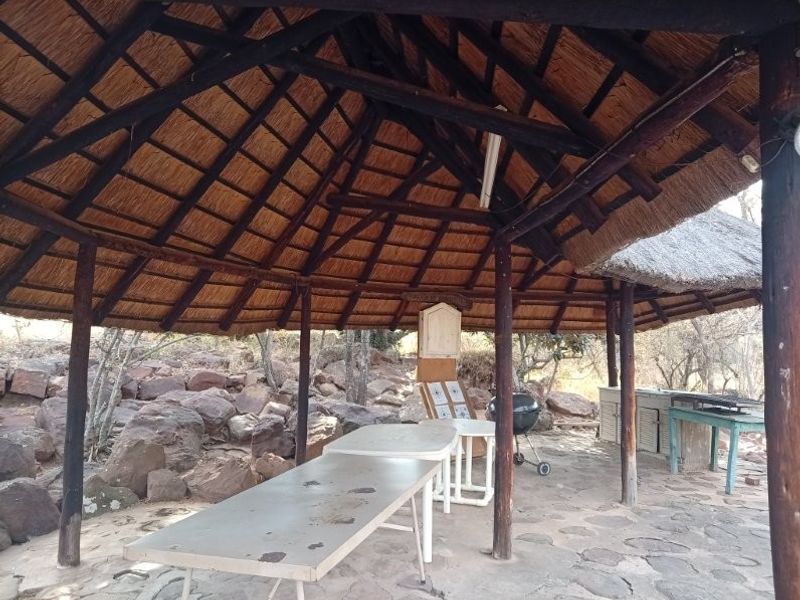 A rustic outdoor thatched lapa area featuring stone paving, timber support structures, and a built-in counter, ideal for outdoor entertaining in a farm setting.