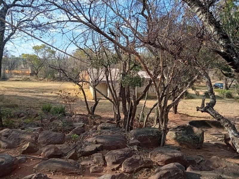 A view of the rear farm grounds featuring natural rocky terrain, mature trees, and a glimpse of the building structure with a metal roof.