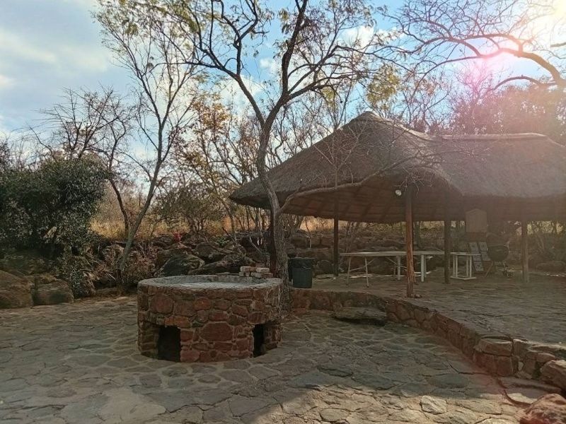 Outdoor entertainment area featuring a rustic thatched lapa, stone-paved flooring, and a built-in stone fire pit set within a natural landscape.