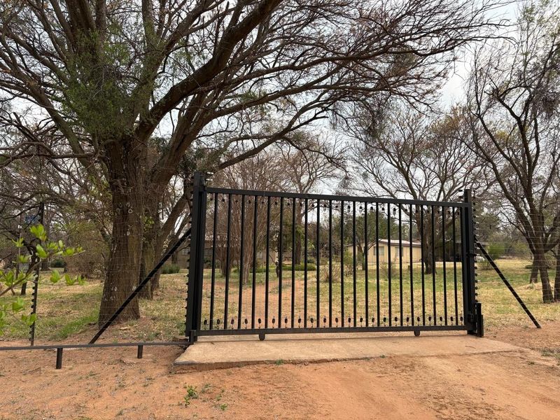 A secure metal sliding entrance gate featuring integrated electric fencing, set within a rural farm property landscape.