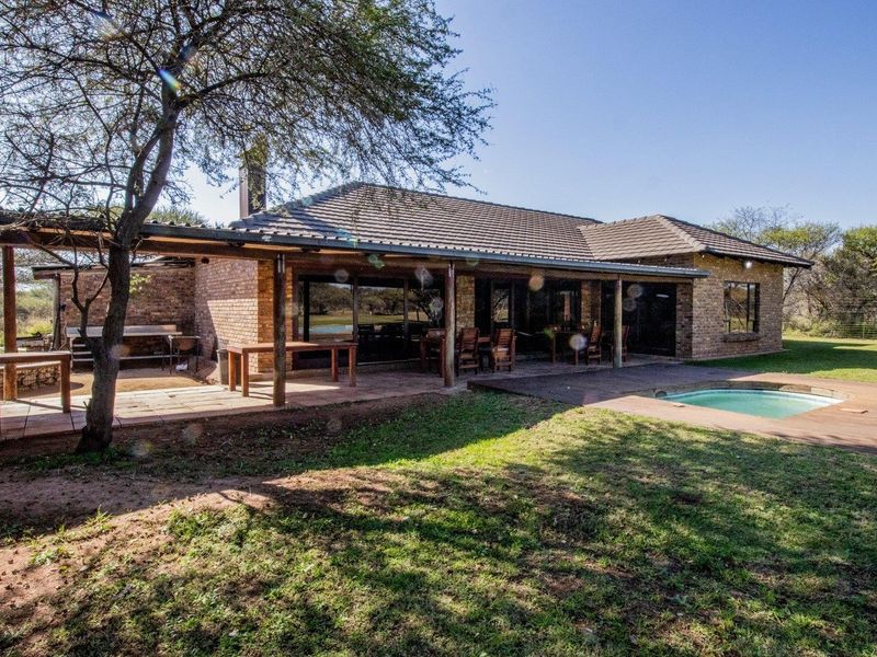 Rear view of a brick farmhouse featuring a covered patio, wooden decking, and a swimming pool set within a spacious garden.