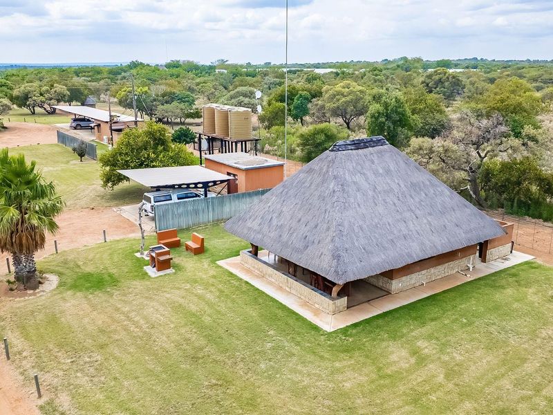 Aerial view of a farm property featuring a main dwelling with a traditional thatch roof, water storage tanks, a carport, and a built-in braai area.