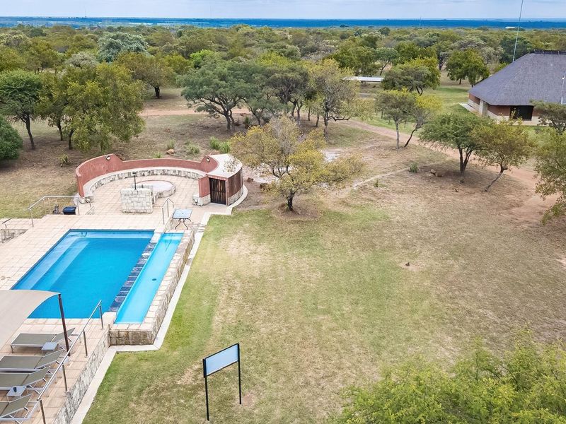 Aerial view of a farm property featuring a swimming pool, built-in braai area, and a large thatched-roof structure set within expansive natural grounds.