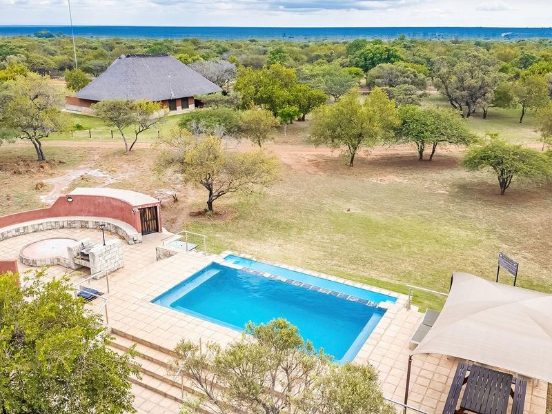 Aerial view of a farm property featuring a swimming pool, a curved masonry braai area, and a separate thatch-roofed building set in a landscape.