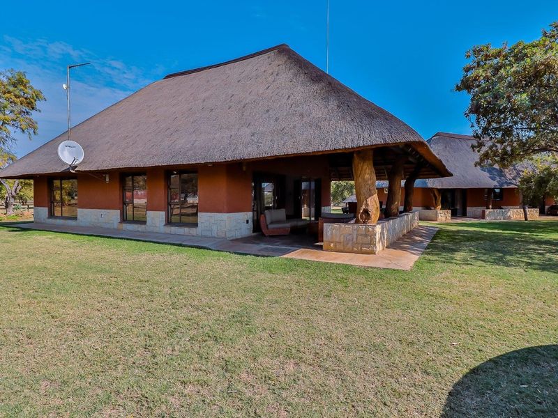 Rear view of a farm-style dwelling featuring a traditional thatch roof, stone-clad base, and a spacious covered patio area set within a landscaped garden.